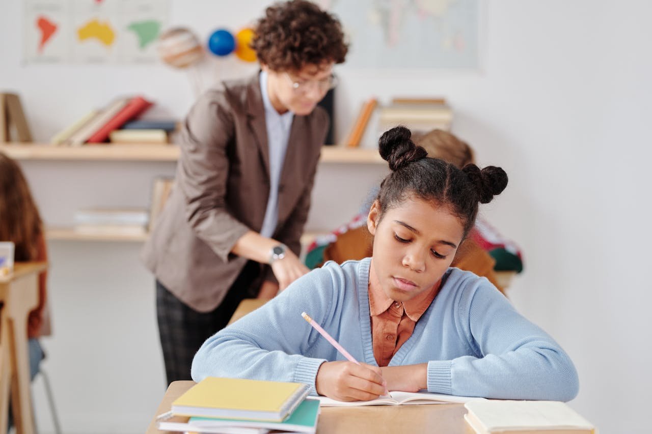 Focused young student writing during a classroom activity with teacher in background.